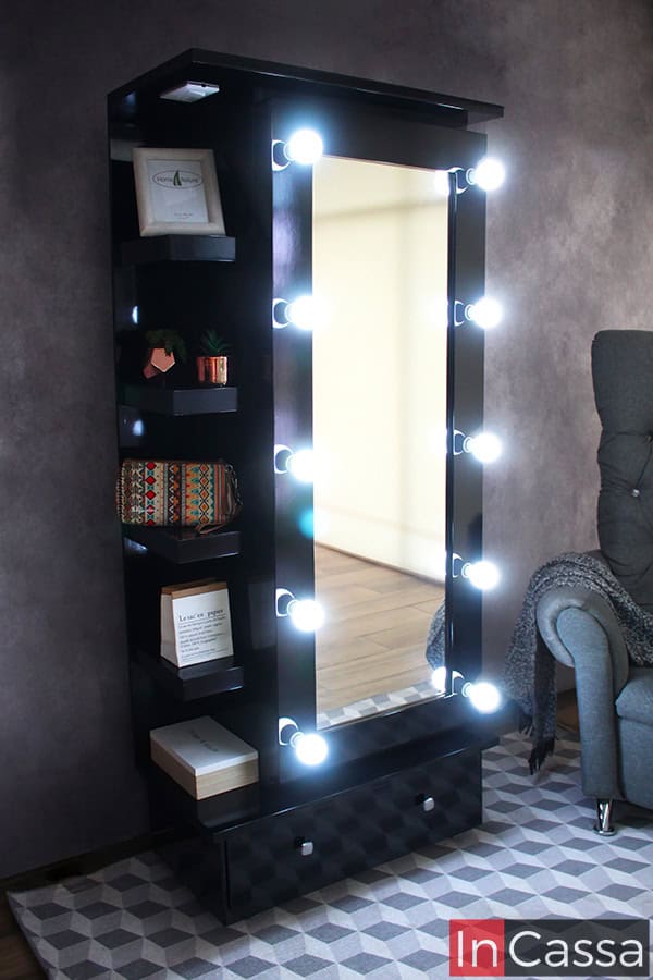 Se presenta la zapatera Piacenza en acabado negro brillante, con espejo de cuerpo completo rodeado por bombillas LED encendidas. El lateral incluye repisas decoradas con libros, plantas y accesorios, mientras que en la parte inferior se integra un cajón amplio con tiradores cuadrados.