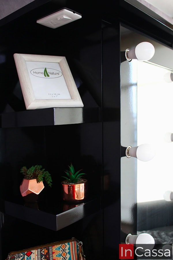 Una toma en primer plano muestra el lateral de la zapatera Piacenza en acabado negro brillante. Se aprecian repisas decorativas con macetas pequeñas de tono cobre y un portarretrato, así como parte del marco con focos LED apagados que rodea el espejo.