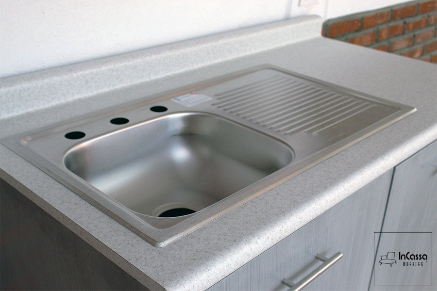 Una vista cercana muestra la tarja de acero inoxidable con escurridor integrada en la cubierta de formaica estilo sal y pimienta. El acabado pulido y los orificios para llaves resaltan la funcionalidad del módulo inferior de la Cocina Integral Modelo PISA para Estufa.