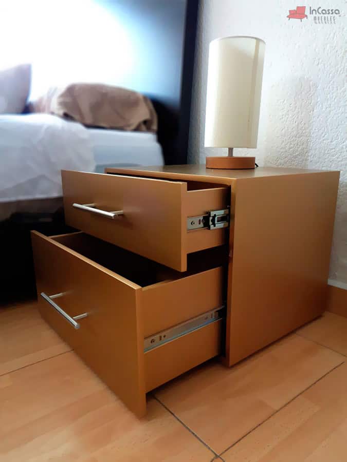 La imagen muestra un ángulo lateral del buró Portugal en tono maple, con ambos cajones abiertos para resaltar su capacidad interna y correderas metálicas. La lámpara decorativa sobre el buró añade un toque elegante al conjunto, junto a la cama parcialmente visible al fondo.