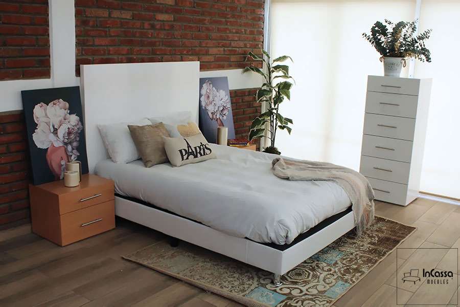 La foto presenta una toma amplia de una recámara decorada con una cabecera blanca, base tapizada, buró en tono maple y cómoda blanca al fondo. La ambientación incluye cuadros florales, plantas decorativas y textiles neutros, generando una atmósfera moderna y acogedora.