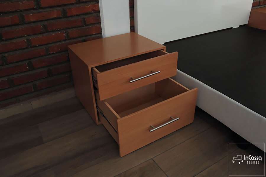 Esta fotografía muestra un buró en tono maple con sus dos cajones parcialmente abiertos, revelando su funcionalidad y espacio de almacenaje. A un costado se aprecia la base tapizada blanca, resaltando la armonía entre los muebles del conjunto.