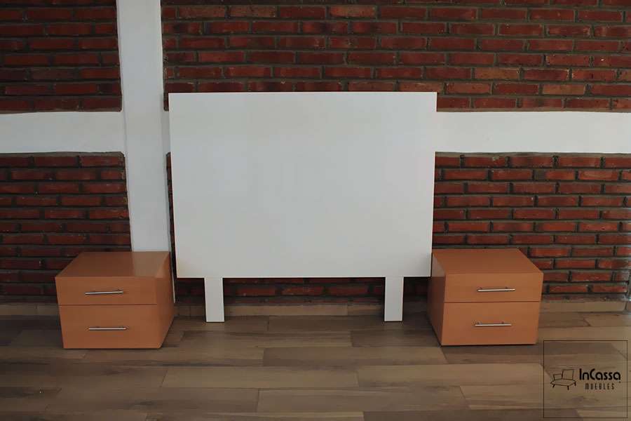 La imagen muestra una vista completamente frontal del juego de recámara conformado por una cabecera blanca de diseño limpio y moderno, acompañada de dos burós en tono maple. Ambos burós incluyen dos cajones con jaladeras metálicas, creando un contraste elegante con el fondo de muro de ladrillo.
