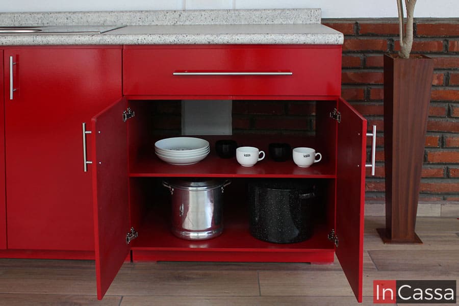 Fotografía de los espacios izquierdos del módulo inferior de la cocina, siendo dos compartimentos divididos por un entrepaño horizontal en los que se apoyan distintos utensilios para mostrar su capacidad. La parte trasera de la estructura se conserva libre, dejando el espacio necesario para la instalación de la parrilla empotrable. La sección completa está pintada en un acabado rojo completo.