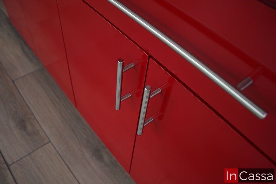 La foto presenta una toma que se concentra en las jaladeras instalas en cada puerta y cajón de la cocina, los cuales son metálicas con diseño de barra "T" en acabado cromado. Consecuentemente, también es posible apreciar más de cerca las puertas instaladas en la cocina, todas de color rojo en un tono brillante.