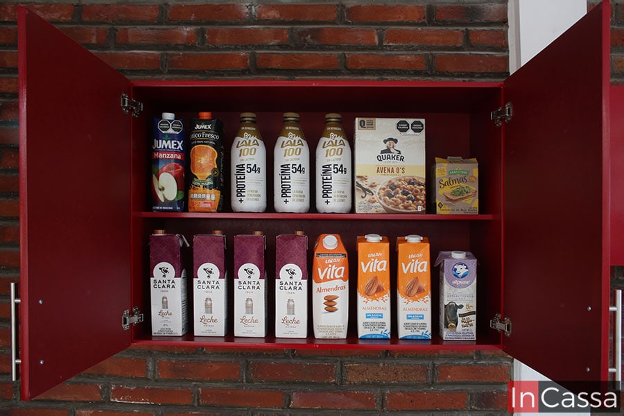 Un vistazo cercano de la alacena superior de la cocina. Ya que se muestran sus puertas abiertas, es posible ver un entrepaño que divide su interior en dos amplias espacios en los cuales se guardan algunos envases y botellas que ejemplifican la capacidad de cada compartimento. El módulo posee un tono rojo en la totalidad de su construcción.