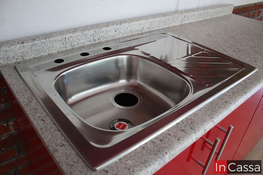 La imagen muestra una toma completa de la tarja de acero inoxidable incorporada a la cubierta de la cocina. La tarja de acabado cromado tiene un diseño de tina sencilla con escurridor del lado derecho, mientras que la cubierta es de formica con un acabado en estilo sal y pimienta.