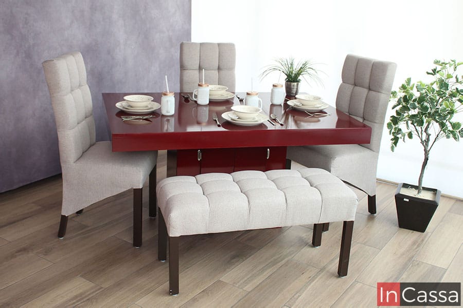 Comedor para 5 personas modelo QUIRICO presentado en una estancia con pared gris y piso de madera, la mesa y pedestal son de color borgoña con acabado brillante, la mesa está acompañada de 3 sillas y una banca, todas tapizadas en lino greige con patas color café.
