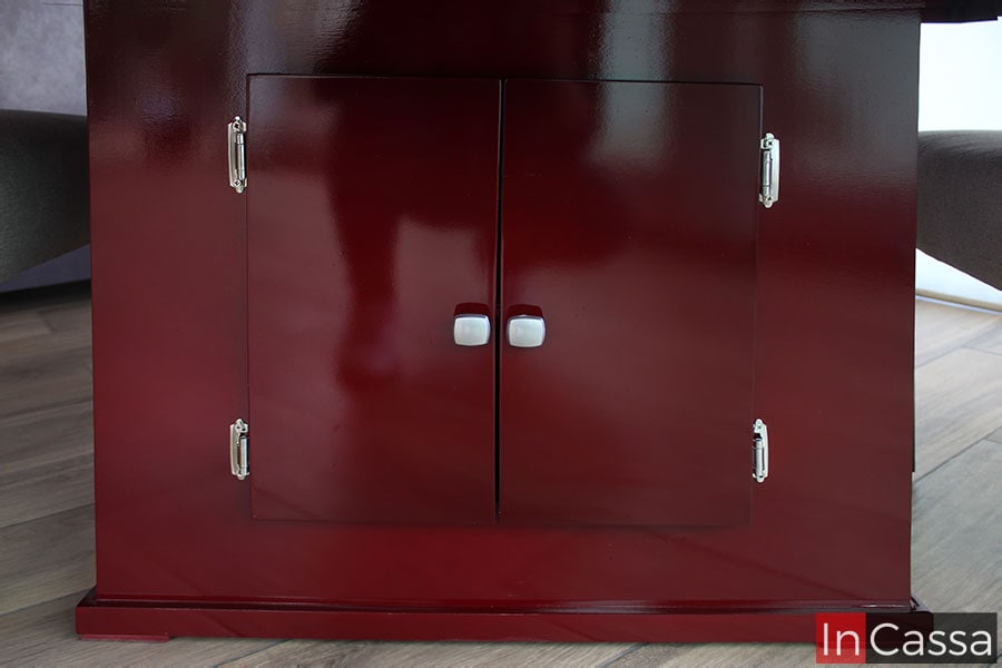 La toma se enfoca completamente en el pedestal, en el cual podemos ver dos puertas que cubren el compartimento junto con parte del sistema de bisagras así como las manijas incorporadas en cada puerta. La estructura completa esta pintada en un tono borgoña con acabado brillante al igual que el resto del mueble.