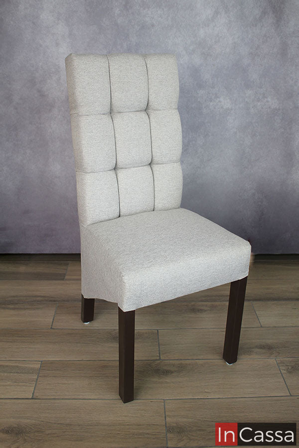 Una imagen completa de una de las sillas que incluye el comedor, presentada en una estancia con una pared gris y piso de madera. Podemos apreciar el tapiz del mueble en lino color greige, además del acabado capitoneado en el respaldo así como sus patas en color café mate.