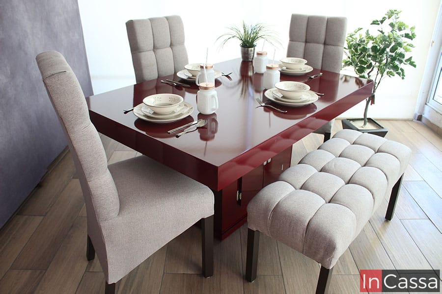 Tenemos una vista lateral del comedor, teniendo una mejor vista del pedestal de la mesa, además de que el acabado brillante de la pintura color borgoña queda mucho más resaltado gracias a la toma. Igualmente podemos notar la banca y una de las sillas más a detalle, sobre todo, en la textura del tapiz lino greige y el acabado café mate en las patas de ambos muebles.