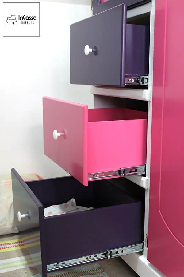 La fotografía muestra una vista lateral del Roperito Infantil Modelo ROMINA, donde se aprecian sus tres cajones abiertos con correderas metálicas de extensión completa. Los frentes en tonos rosa pastel y uva aportan un estilo moderno y alegre, mientras que la estructura blanca resalta la calidad y el acabado del mueble.