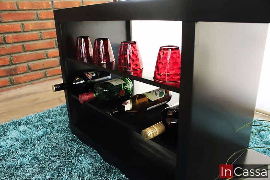 Se presenta una vista lateral en ángulo bajo del módulo de almacenamiento de la cantina ROSWELL, donde se exhiben botellas y vasos rojos sobre entrepaños abiertos. El diseño moderno en acabado chocolate resalta sobre la alfombra azul y el fondo de ladrillo, brindando estilo y funcionalidad.