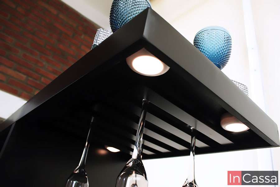 En esta toma en ángulo contrapicado se aprecia el elegante portacopas de la cantina ROSWELL, fabricado en acabado chocolate con luz LED empotrada. Las copas suspendidas y la iluminación crean un efecto visual sofisticado y funcional.