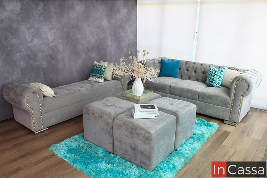 La imagen muestra una sala moderna en estilo glam tapizada en suede gris perla compuesta por un sofá capitoné, un diván con detalles en tachas doradas y 4 taburetes. Los cojines en tonos vibrantes y la alfombra azul contrastan, aportando frescura y dinamismo al espacio.