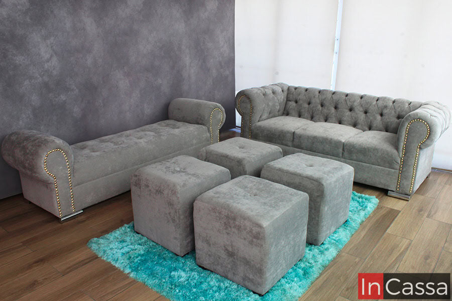 La imagen muestra un conjunto de sala, tapizado en suede gris perla, con sofá capitoneado, diván y cuatro taburetes que forman una mesa central. La alfombra azul brillante contrasta y añade un toque de color vibrante, logrando un ambiente cálido y acogedor.