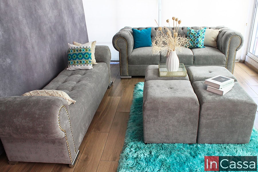 Se presenta una sala elegante tapizada en suede gris perla con un sofá y un diván capitoné, embellecidos con tachas doradas en los brazos, además de 4 taburetes. La alfombra turquesa y los cojines en tonos azul y verde añaden color y vida al ambiente.