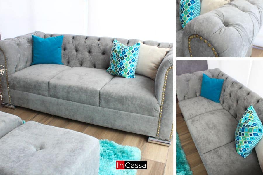 La imagen muestra un sofá de suede gris perla capitoneado, de estilo glam, con tachas doradas en los brazos y patas metálicas. Los cojines decorativos en tonos azules y patrones geométricos aportan un toque moderno y atractivo al diseño.
