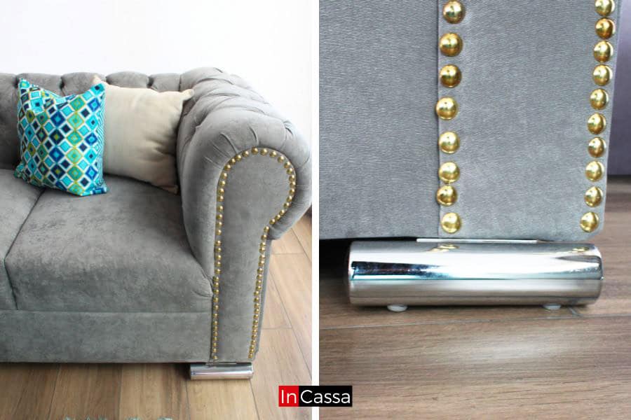 La imagen muestra el brazo de un sofá tapizado en suede gris perla con capitoné y tachas doradas, combinando elegancia y durabilidad. La base metálica le aporta un toque moderno y estable, mientras que los cojines en tonos vivos añaden contraste y confort.