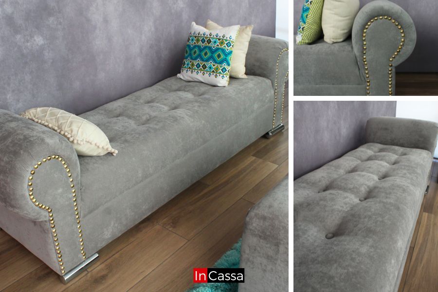 La foto presenta un diván elegante tapizado en suede gris perla con acabado capitoné y detalles de tachas doradas que destacan su diseño clásico. Los cojines en tonos claros y vibrantes crean un contraste que ilumina el ambiente.
