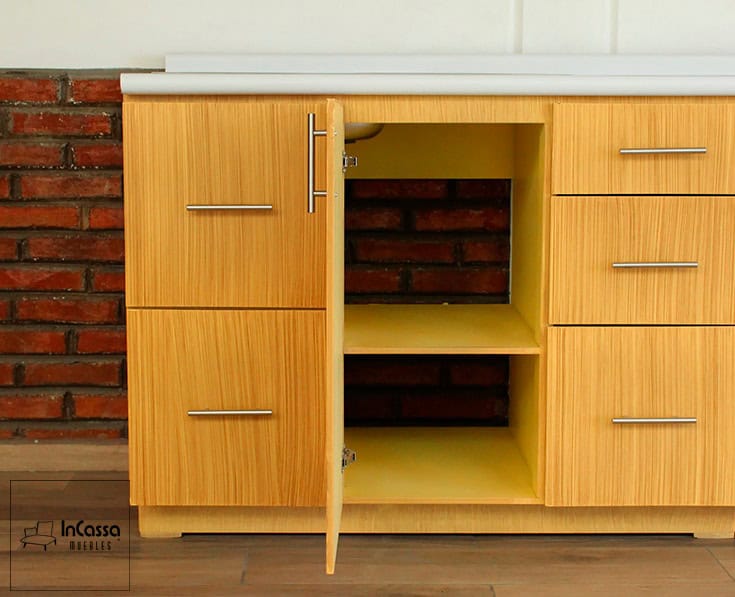 En esta toma frontal del módulo inferior destaca una de sus secciones, el cuál se presenta con su puerta abierta dejando ver dos compartimentos divididos por un entrepaño, todas las partes de esta sección son de un tono natural parecido al laqueado presente en las partes exteriores. Adicionalmente, está sección se fabrica con espacio hacia la pared, para la instalación de la plomería para la tarja.