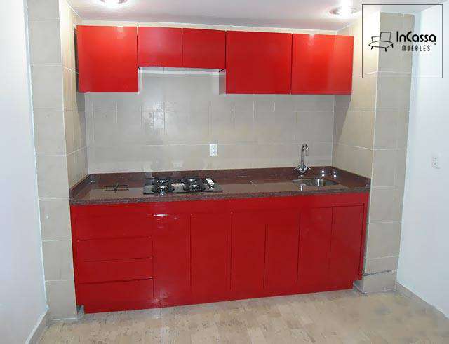 Se presenta una cocina integral en acabado rojo vivo brillante con diseño minimalista, cubierta de formaica en estilo mármol negro y tarja de acero inoxidable. La imagen frontal permite apreciar tanto los módulos superiores como inferiores, con espacios para parrilla y campana.