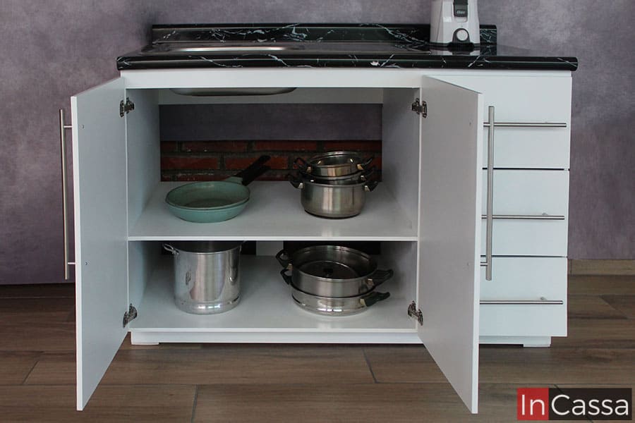 La foto se concentra en el interior del modulo de tarja, en el cuál podemos observar un acabado en color blanco en la estructura interna así como en el entrepaño, en el cuál se apoyan distintos utensilios de cocina ejemplificando la capacidad del mismo. Esta parte queda hueca, ya que es necesario el acceso a la pared para la instalación de la plomería.