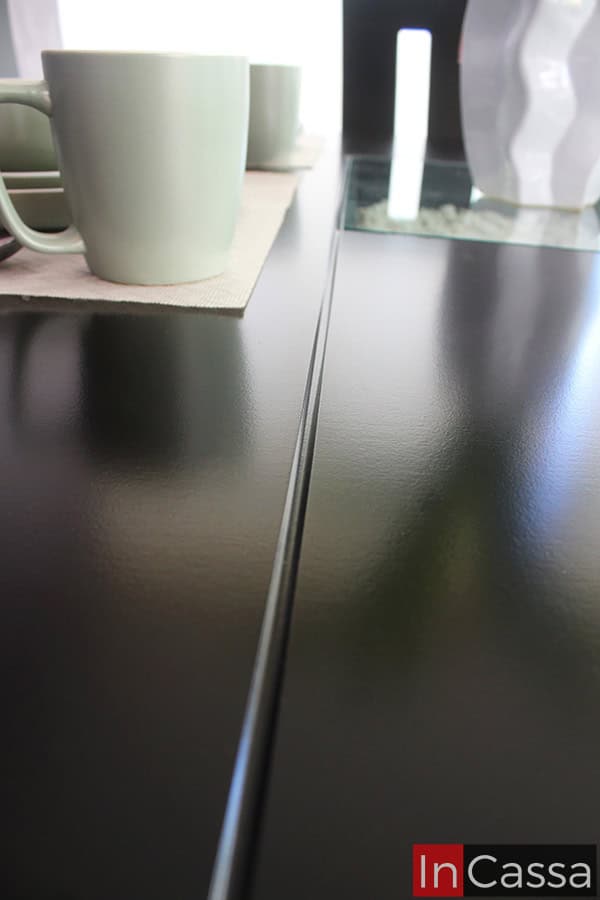 La foto resalta la textura lisa y el brillo del acabado en tono chocolate del comedor. Se aprecia con claridad el fino biselado de las uniones, así como el elegante arenero con decoración central, lo que aporta un toque contemporáneo y distinguido.