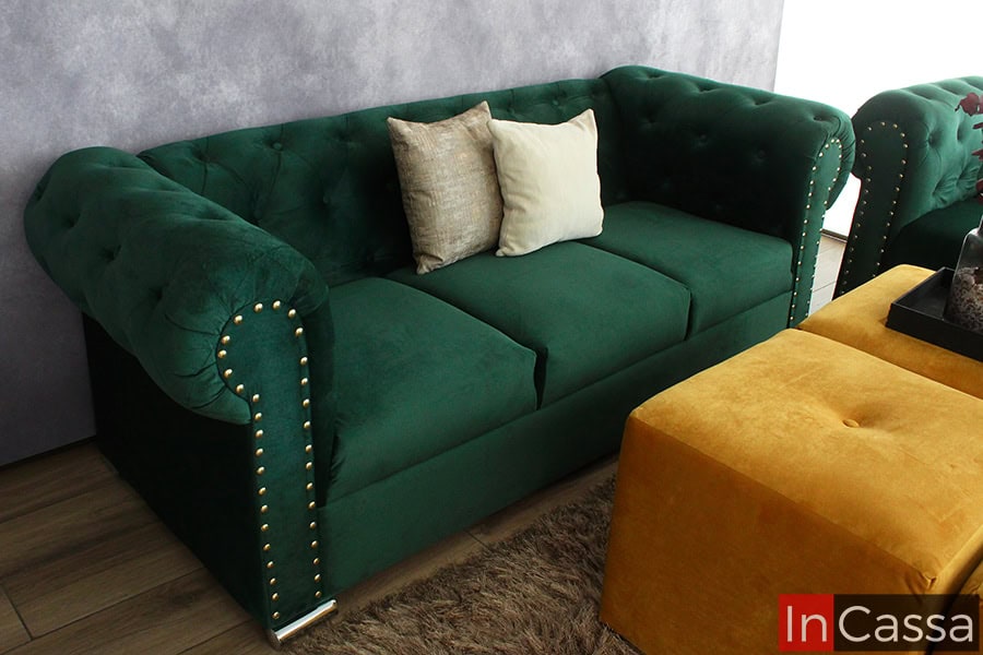 La imagen muestra el sofá del modelo San Petersburgo en tapizado suede verde botella, con acabado capitoneado, remaches dorados y cojines decorativos. Sus patas cromadas aportan un toque elegante al diseño clásico.