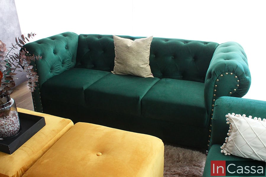 La foto presenta el sofá del modelo San Petersburgo desde una toma frontal tapizado en suede verde botella. Destacan el diseño capitoneado, remaches dorados y cojines decorativos, combinados con taburetes dorados al centro para un estilo sofisticado.