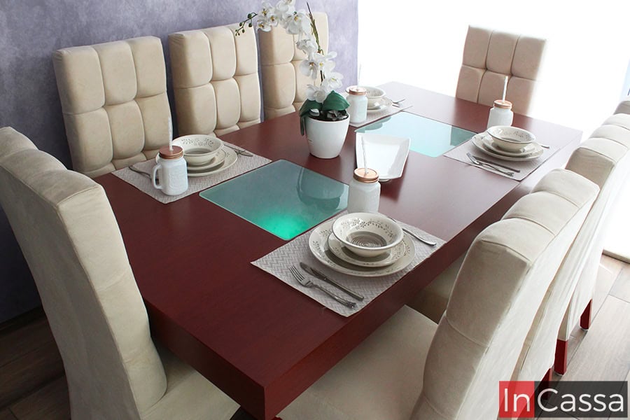 Toma frontal que exhibe el comedor Seúl rectangular con cubierta tabaco y dos paneles de vidrio esmerilado al centro. Las sillas altas, tapizadas en suede hueso con capitonado en el respaldo, enmarcan la mesa, complementada con vajilla blanca y detalles decorativos sobre un piso estilo madera.