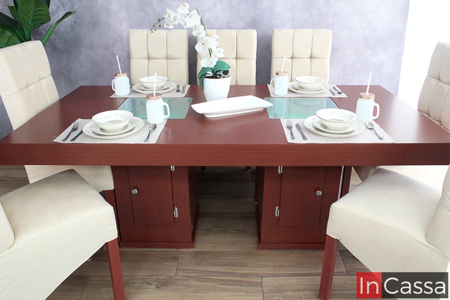 La foto presenta la mesa rectangular en tono tabaco con paneles centrales de vidrio esmerilado, sostenida por dos pedestales con puertas y herrajes metálicos. Está acompañada por sillas altas tapizadas en suede hueso capitonado, sobre un piso de acabado tipo madera.