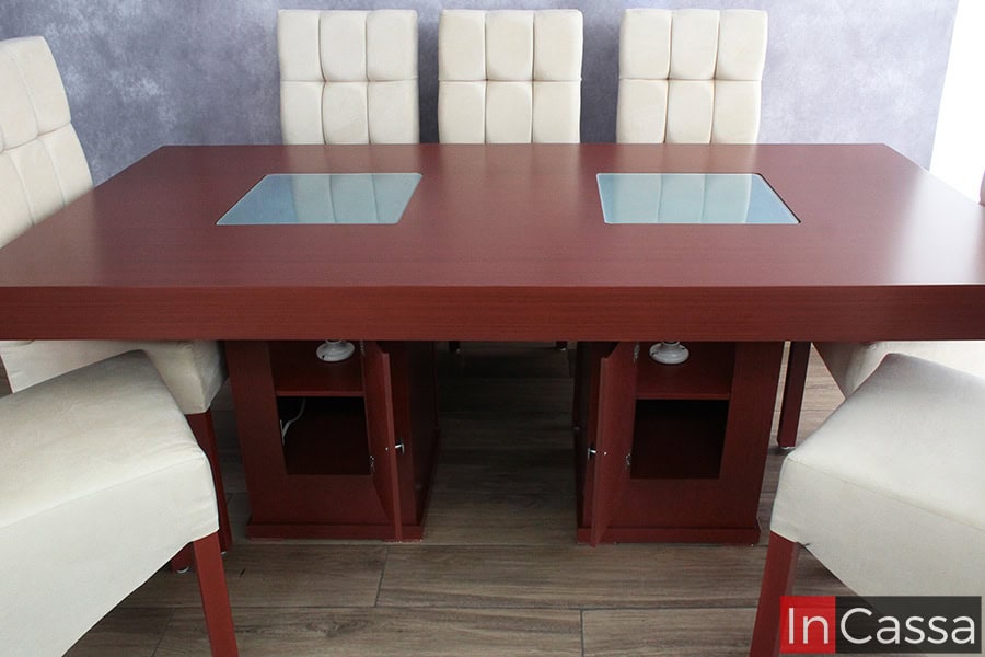 Se presenta una vista frontal del comedor Seúl para ocho personas en acabado tabaco, mostrando los pedestales con puertas abiertas que revelan compartimentos internos y luminarias para la iluminación LED de los paneles de vidrio esmerilado. Las sillas altas tapizadas en suede hueso capitonado completan el conjunto sobre piso tipo madera.