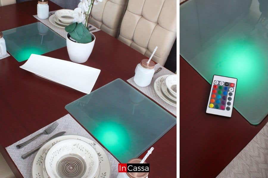 La imagen muestra la superficie de la mesa en tono tabaco con paneles de vidrio esmerilado iluminados con luz LED verde controlable por mando a distancia. La mesa está decorada con vajilla, cubiertos y un florero central, acompañada de sillas tapizadas en suede hueso capitonado.