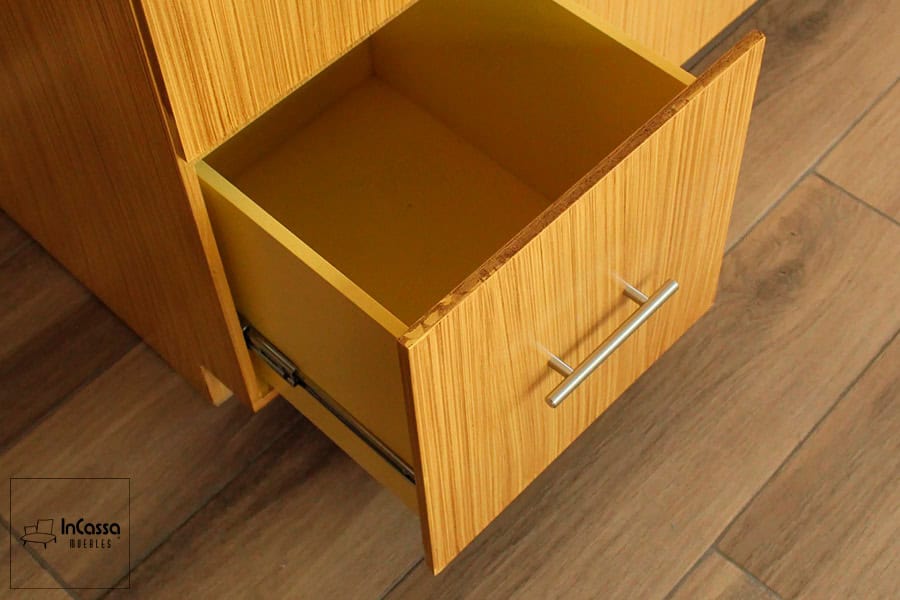 Se nos enseña uno de los cajones del módulo inferior abierto, dejando a la vista su estructura de acabado natural completa. en la que se ofrece una vista cercana de detalles tales como son: uno de sus rieles metálicos y una jaladera de barra T, ambos parte de su sistema de apertura y cierre. El cajón cuenta con un interior amplio y con buena profundidad.