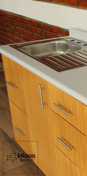 Tenemos una vista lateral del módulo inferior, desde la que podemos apreciar de mejor manera el acabado laqueado natural de su estructura, al igual que parte de su cubierta de formaica blanca y su tarja de acero inoxidable. Además, también se pueden observar las jalades de barra T incorporadas a los cajones y puertas del módulo.