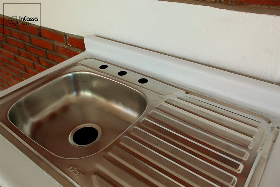 Foto de la tarja de acero inoxidable que cuenta con una tina en su lado izquierdo y un escurridor en el derecho. La toma también da una vista parcial pero cercana de la cubierta de formaica de color blanco.