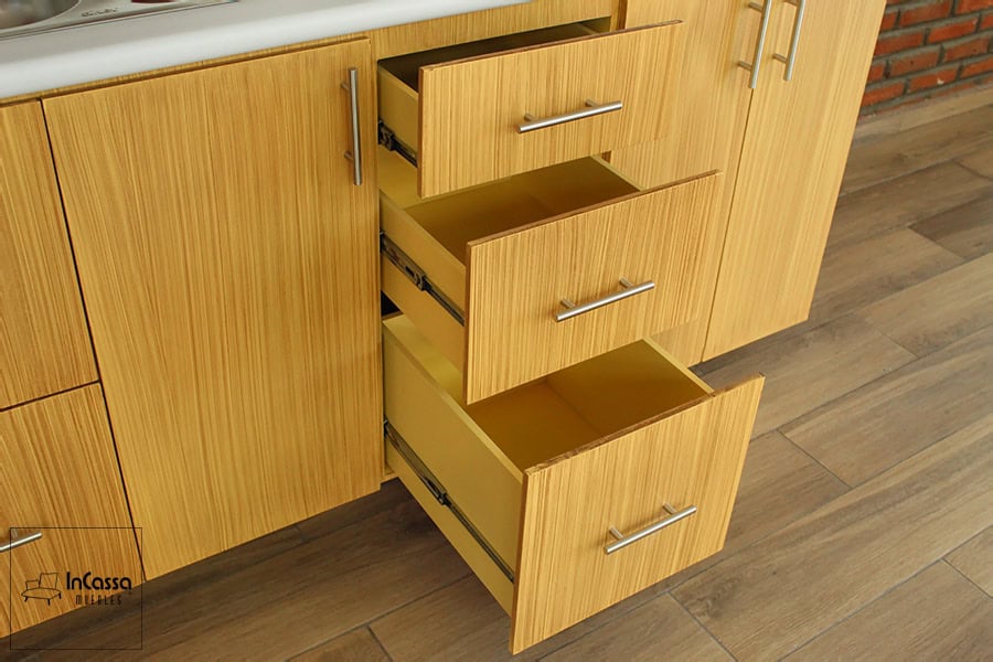En esta foto se presenta buena parte del módulo inferior de acabado laqueado natural, en la que destacan tres de los cajones que incluye el módulo, presentándose abiertos dejando apreciar su estructura completa, además de sus rieles metálicos y sus jaladeras de barra T.