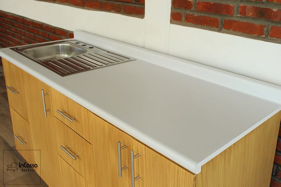 En está toma destaca en su totalidad la cubierta del módulo inferior, dejándonos observar detalladamente su acabado blanco e incluso la tarja de acero inoxidable incluida e instalada en la cocina. Además, también ofrece una vista parcial de los cajones y puertas, en los que podemos apreciar su acabado laqueado natural, y algunos detalles adicionales como son sus jaladeras de barra T.