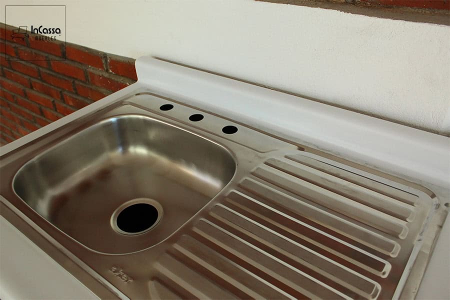 La imagen destaca la tarja integrada a la cubierta blanca de la cocina. Está es de acero inoxidable, lo que le da un acabado cromado, con tina del lado izquierda y escurridor del derecho.