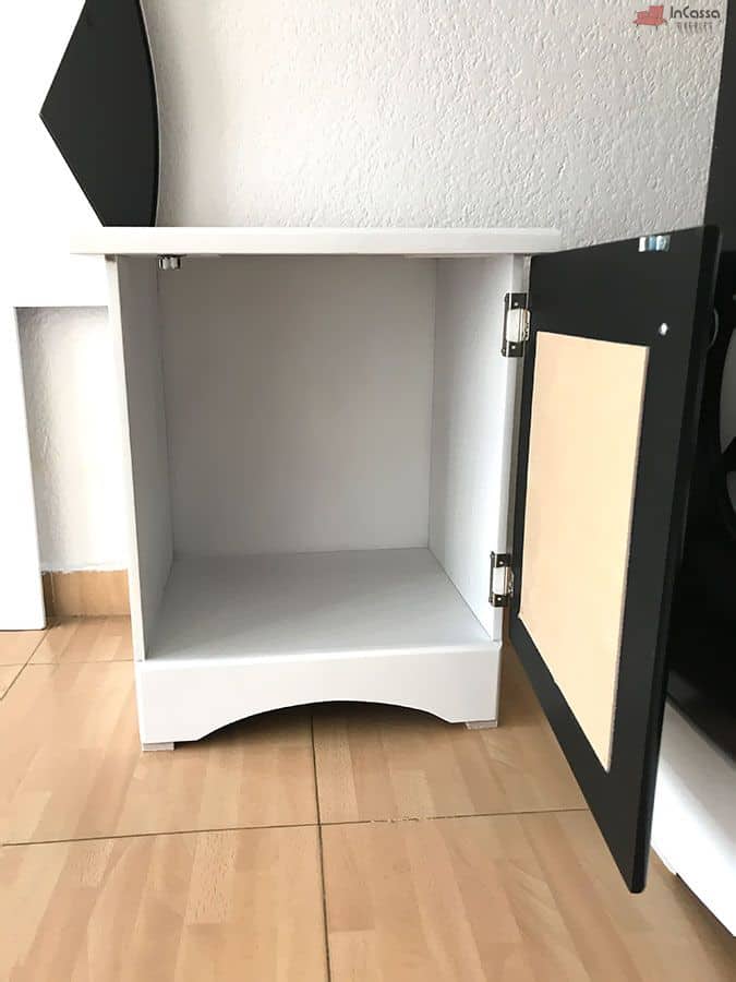 La imagen muestra el buró blanco con puerta negra abierta, revelando un amplio espacio interior sin divisiones para almacenamiento. Sus herrajes metálicos y base con diseño curvo ofrecen practicidad y estilo deportivo.