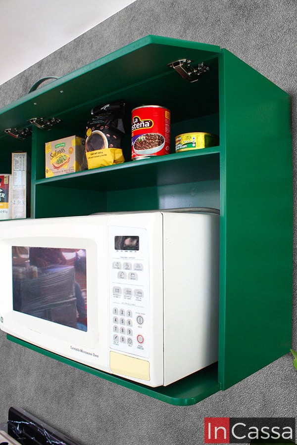 Fotografía desde una toma lateral de la alacena superior de la cocina, concentrándose en el espacio para microondas del módulo, además de su pequeño compartimento adicional para almacenar alimentos enlatados o utensilios. La totalidad del módulo presenta un color verde oscuro en acabado brillante.