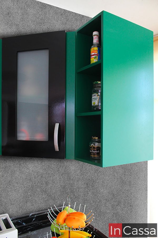 Foto desde una toma lateral del módulo superior esquinero de la cocina, cuya estructura completa es de color verde con excepción de su puerta, la cual es de cristal con marco de MDF negro. También pueden apreciarse 3 de sus estantes abiertos, los cuales son bastante espaciosos y permiten almacenar especias y consumibles de cocina.