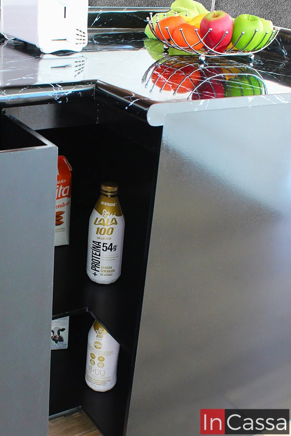 Fotografía del modulo esquinero inferior de la cocina, el cuál es presentado con su puerta con forma de L abierta, mostrando sus dos compartimentos internos los cuales son lo suficientemente amplios para almacenar consumibles o utensilios de cocina. La totalidad del mueble mantiene el mismo tono negro, combinando a la perfección con su cubierta de formaica de estilo de mármol negro.