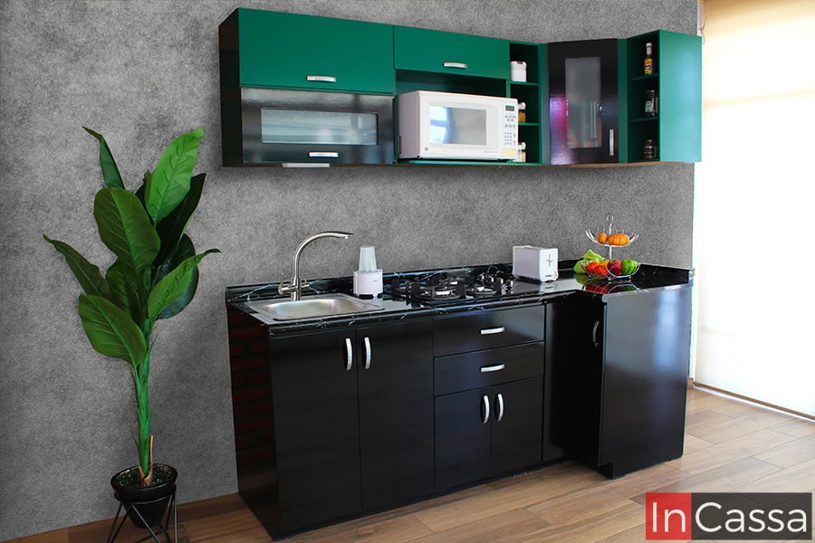 Practica cocina en escuadra de 2.10m x 80cm. La cocina esta conformada con dos módulos inferiores negros y dos módulos superiores de color verde, entre los que destaca su alacena superior con espacio para microondas, además de que todos los módulos inferiores cuentan con cubierta de formaica en estilo mármol negro. La cocina se encuentra instalada en una habitación con paredes de cemento pulido con piso de madera.
