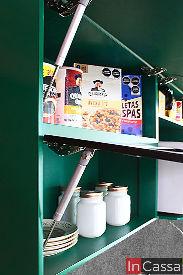 La imagen muestra la alacena superior de la cocina con sus puertas abiertas, estas cuentan con pistones neumáticos para su apertura y cierre de manera vertical. Como muestra la foto, la alacena cuenta con 3 amplios compartimentos para almacenar vajillas, utensilios y alimentos empaquetados o enlatados; cada compartimento mantiene en su interior el mismo color verde que puede verse en sus partes exteriores.