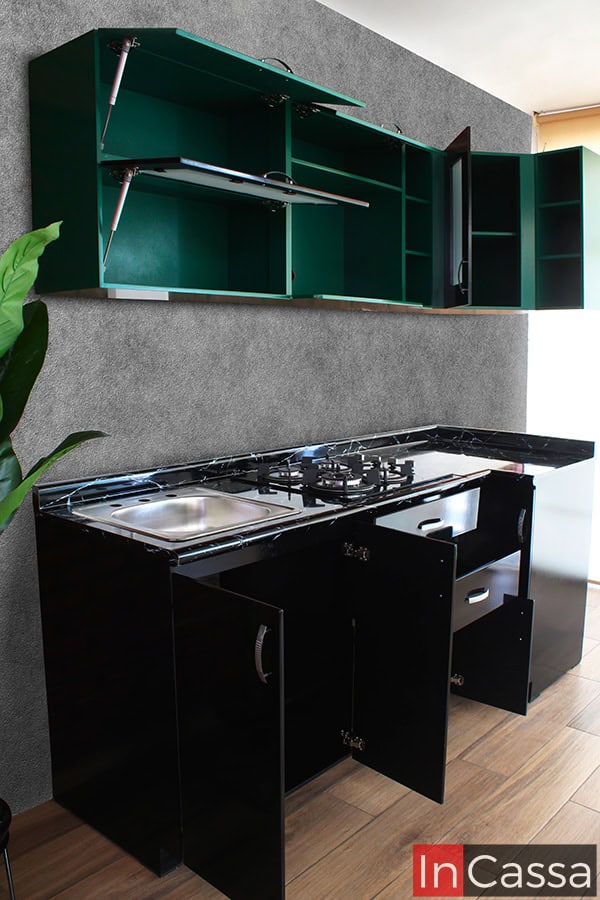 Una toma lateral de la cocina completa, la cuál ofrece una vista más completa de todos sus módulos, los cuales se presentan con sus puertas y cajones abiertos, permitiendo apreciar sus interiores. La cocina presenta una combinación minimalista de colores, siendo sus módulos superiores de color verde completo, mientras que los módulos inferiores son de color negro en su totalidad, estos últimos además cuentan con cubierta de formaica negra y una tarja de acero inoxidable.