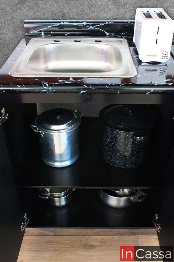 Una foto de la sección de tarja del módulo inferior de la cocina, el cuál se presenta con sus puertas abiertas permitiendo observar sus dos compartimentos internos; estos están divididos por un entrepaño horizontal y ofrecen un amplio espacio para almacenar recipientes y utensilios de cocina. Tanto las partes internas como externas del módulo mantienen el mismo tono negro.