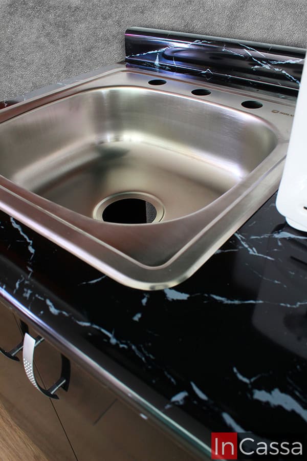 Esta toma se concentra en la tarja de acero inoxidable incluida en la cocina, la cual cuenta con una tina sencilla y sus respectivos espacios para la instalación de llaves mezcladoras. La foto además también proporciona una vista más cercana de la cubierta de formaica del módulo inferior, la cuál es de color negro con acabado brillante.