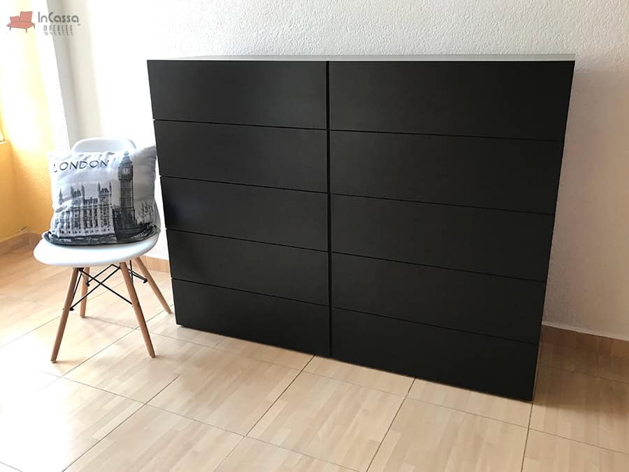 En esta imagen se aprecia una vista frontal de la cajonera Tel Aviv completamente cerrada, en color chocolate. Está colocada junto a una silla blanca con cojines decorativos, sobre un piso laminado claro y junto a una ventana que permite la entrada de luz natural.